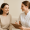 Smiling woman in a beige sweater sits and talks with a female healthcare or wellness professional holding a clipboard.
